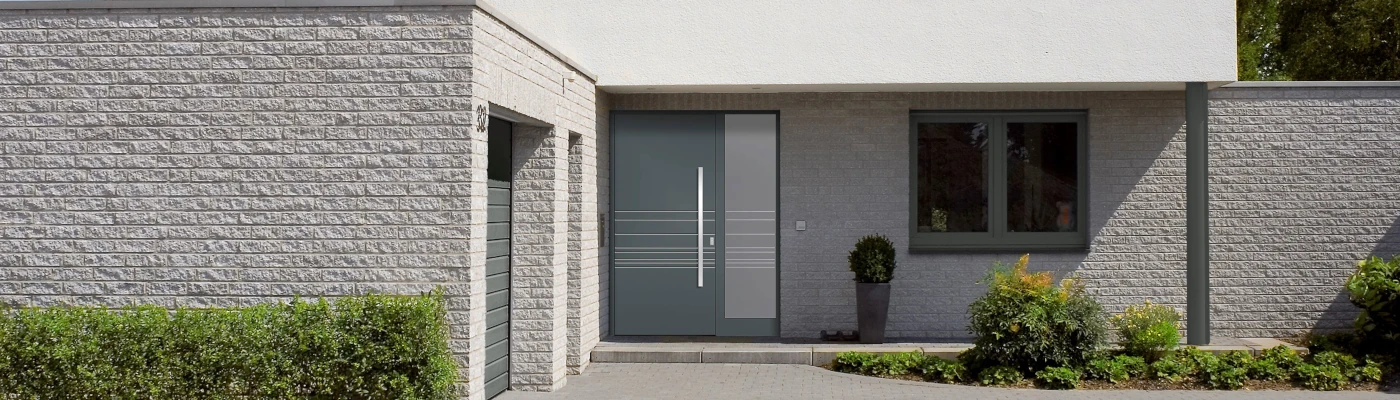 Modernes Wohnhaus mit heller Klinkerfassade, flachem Eingangsbereich und großer grauer Haustür mit langem Griff; daneben ein Fenster sowie gepflegte Sträucher und Pflanzen im Vorgarten.